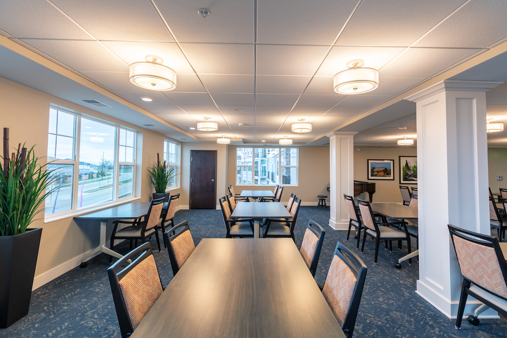 A brightly lit room with several tables, chairs, windows, and potted plants.