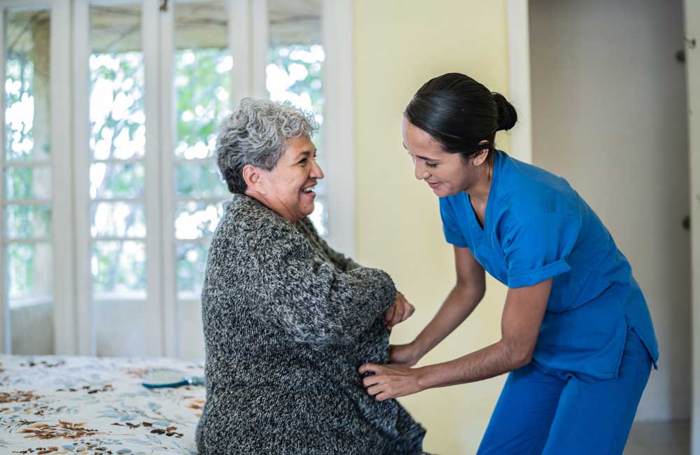 senior-grooming Caregiver assisting an elderly woman with her sweater while both are smiling warmly at each other.