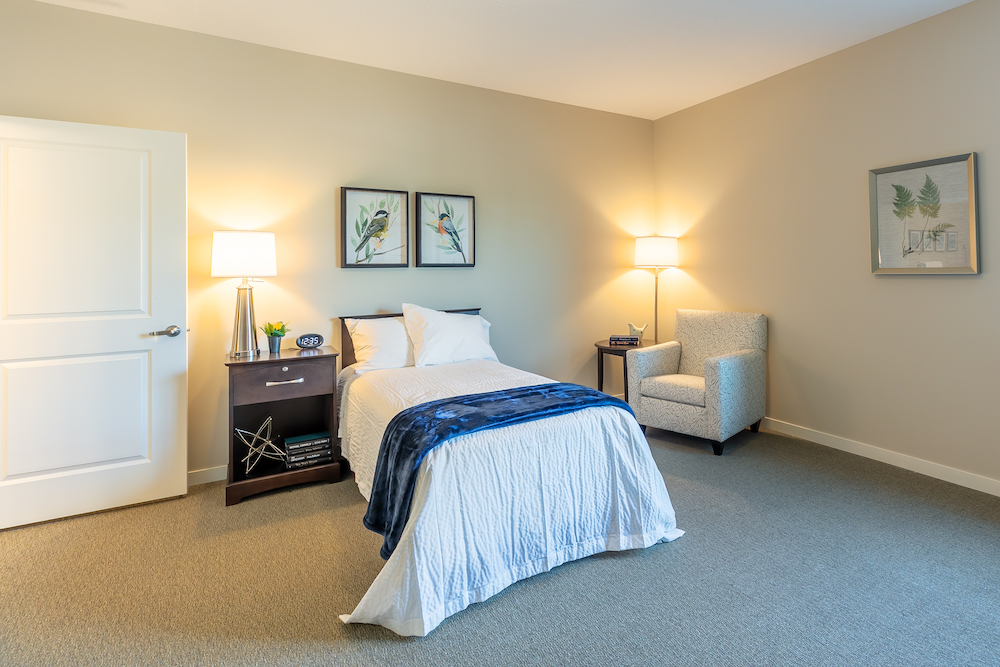 A neatly furnished bedroom with a single bed, two lamps, a cozy chair, and decorative wall art.