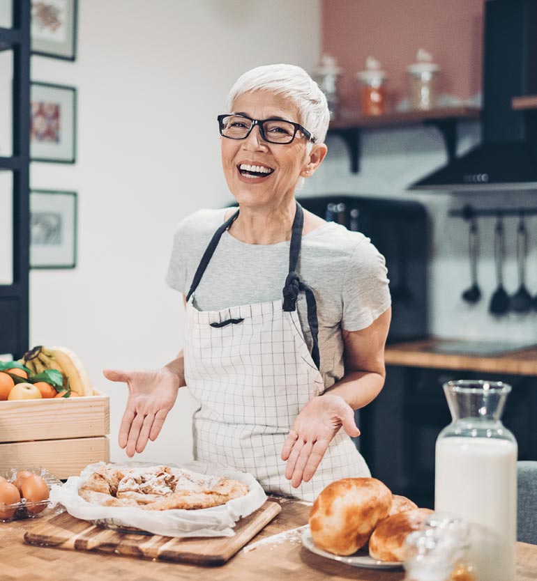 senior-baking Elderly woman in a checkered apron smiling in a kitchen, displaying freshly baked pastry and bread.