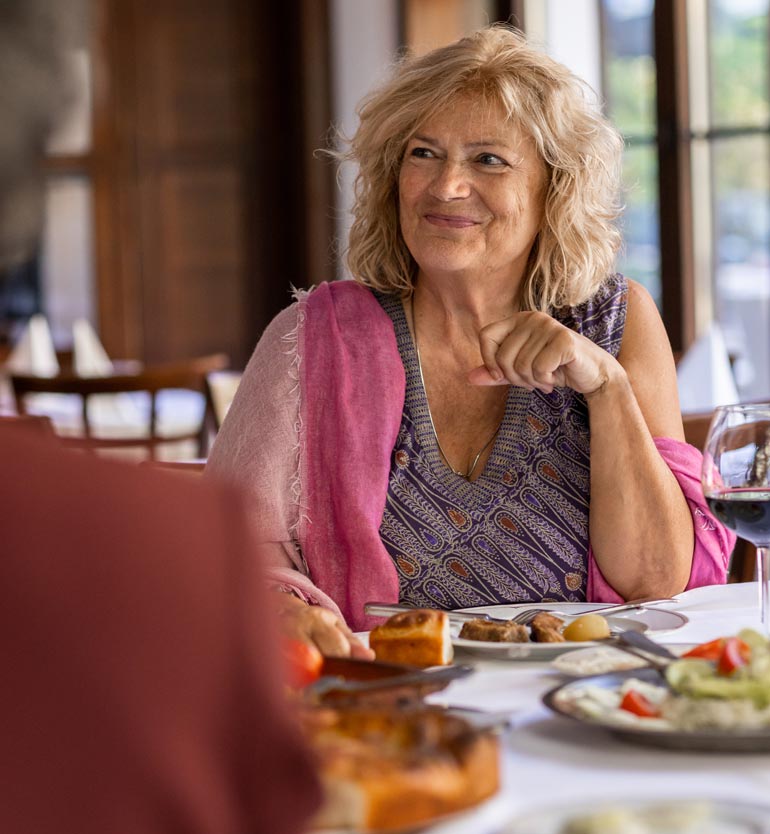 senior-friends-eating Elderly woman enjoying a meal with friends at a restaurant, smiling and looking content.