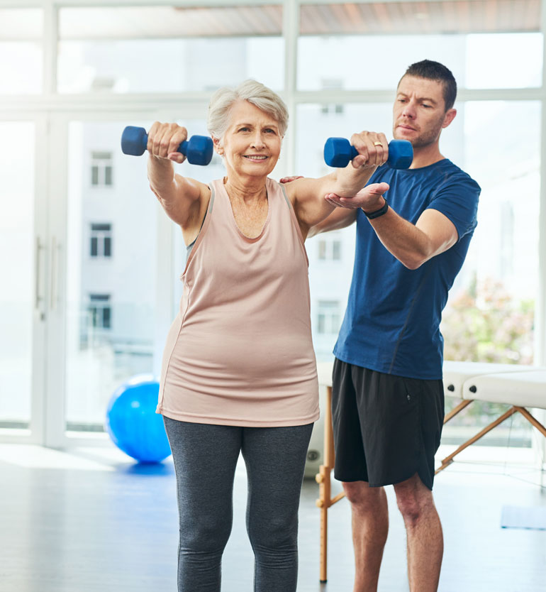 senior-lifting Elderly woman lifting dumbbells with assistance from a trainer in a bright gym setting.