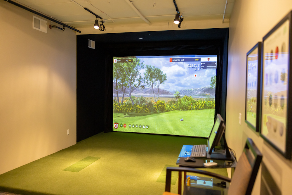A golf simulation room with a green turf floor and a large screen displaying a virtual golf course.