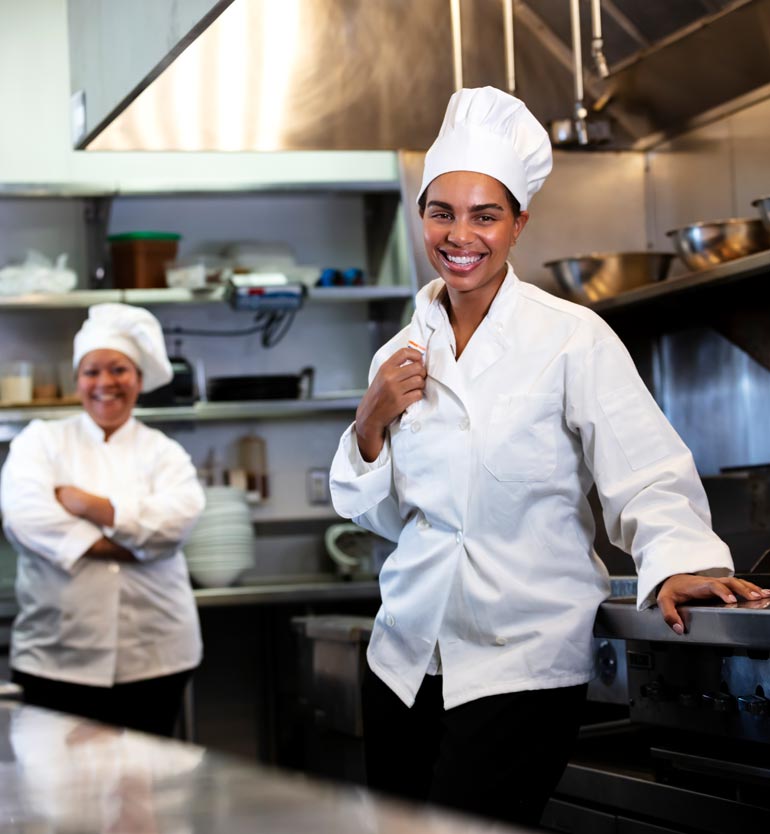 senior-living-chef Two cheerful chefs in a professional kitchen, one smiling at the camera and another standing in the background.