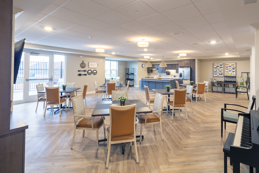 Spacious dining area in a modern senior living facility with tables, chairs, and open kitchen.