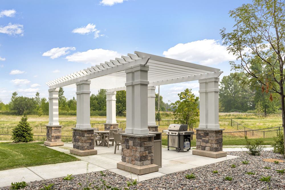 Pergola with stone pillars and outdoor furniture beside manicured lawn and grassy field, under blue sky.