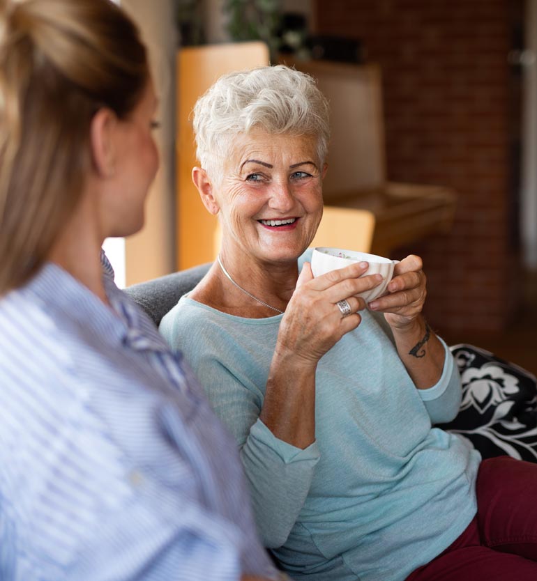 senior-with-team-member Older woman smiling while holding a cup of tea, conversing with a younger woman in a relaxed setting.