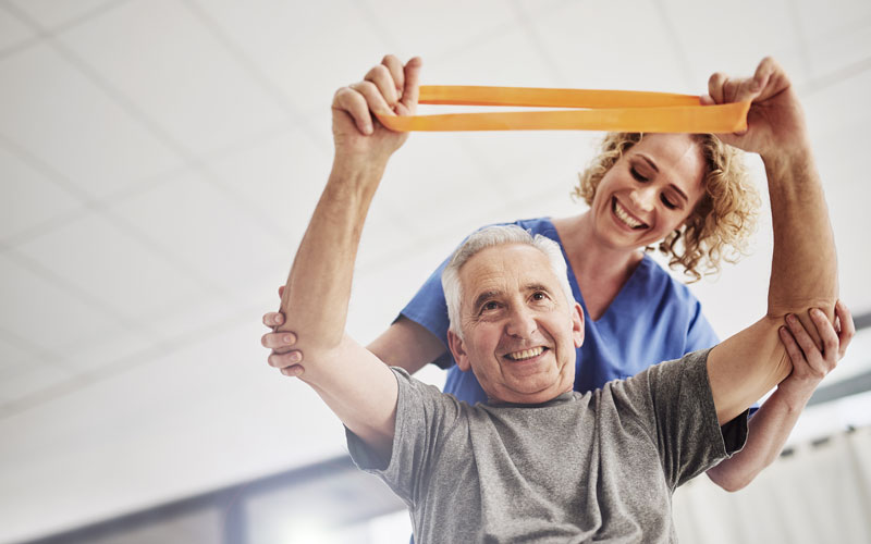 senior-wellness Senior man exercising with resistance band assisted by a smiling caregiver in a bright room.