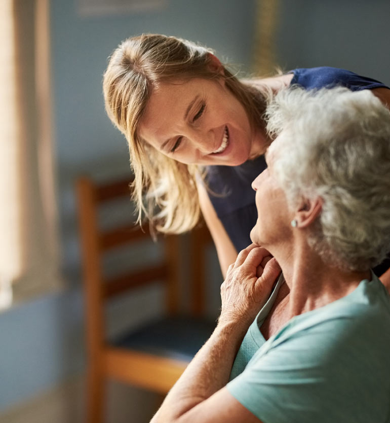 _-senior-memory-care-caregiver- Caregiver smiling at elderly woman in a cozy room, fostering a compassionate environment.