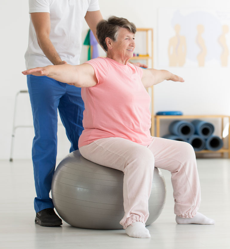 senior-therapy-session A senior woman doing arm exercises on a stability ball during a therapy session with a therapists support.