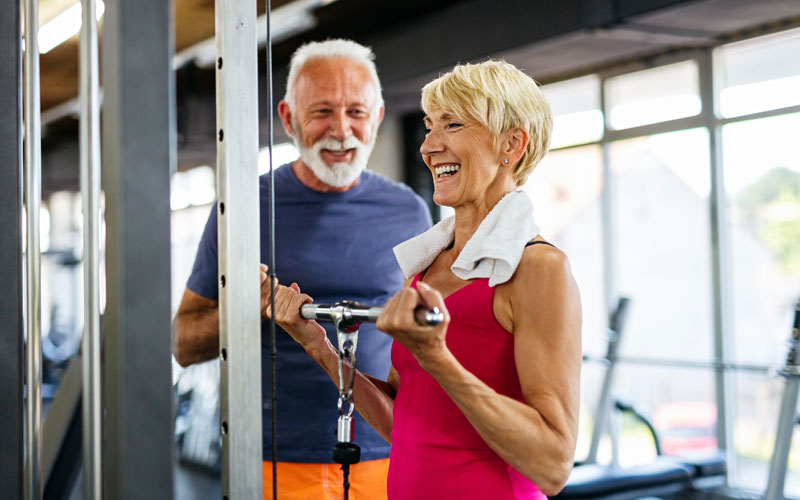 seniors-in-gym Two happy seniors exercising together in a gym, focusing on strength training with gym equipment.