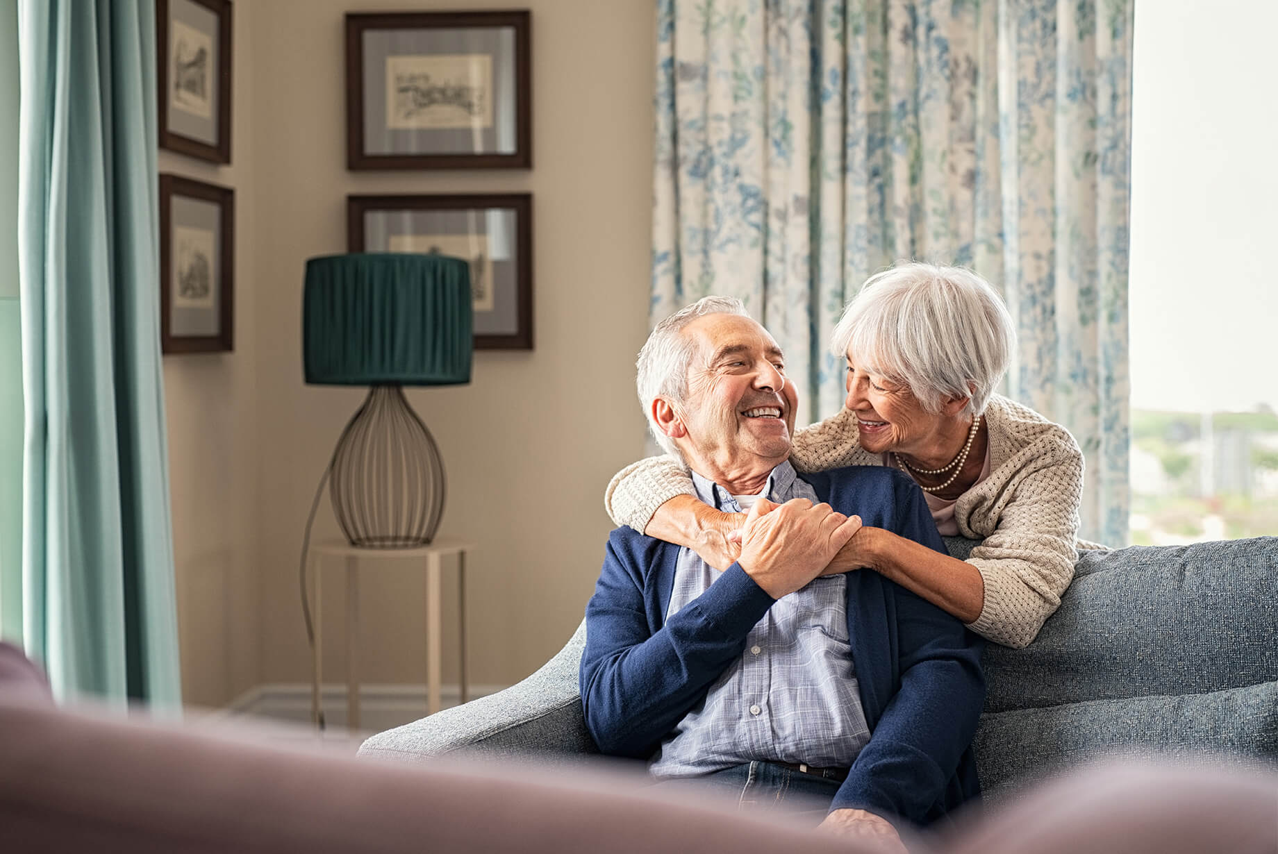 senior-couple-embrace-indoor-on-couch Senior woman stands behind couch and embraces senior man while they smile at each other